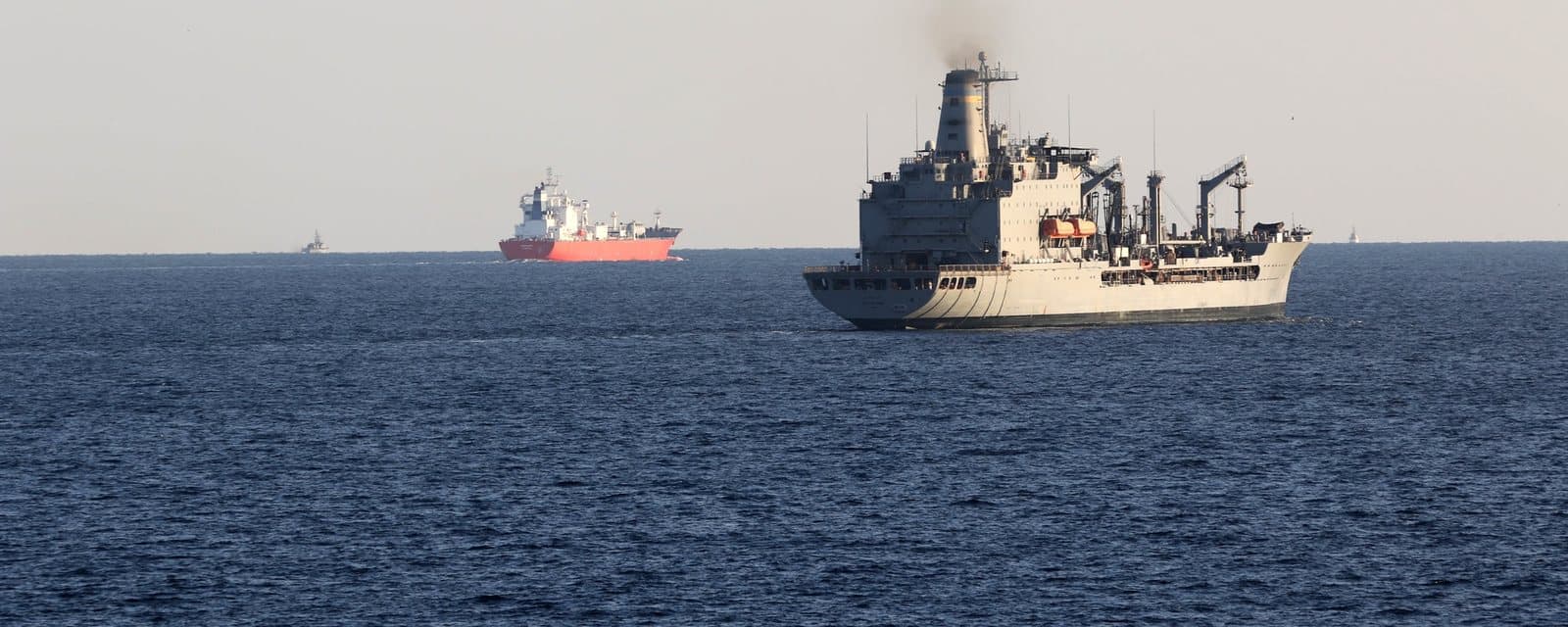 A grey Military Sealift Command oiler at sea, with a red-hulled tanker beyond it, in the Strait of Hormuz